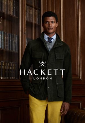 Donkergroene suède jas met meerdere zakken, gedragen over een grijze gebreide trui en een gestreept blauw overhemd, gecombineerd met gele broek.