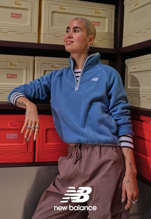 Lichtblauwe fleece-trui met een hoge hals en een halve rits, gecombineerd met losse beige broek. Accessoires omvatten meerdere ringen en groene nagels.