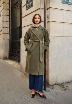 Olivgrön ullkappa med bälte, stora slag och sidfickor, bärs över vida denimbyxor och spetsiga svarta ballerina.