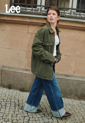 Veste oversize vert olive, top blanc, jean bleu à jambes larges avec ourlets retroussés, bottes à talons marron foncé, debout sur une rue en pavés.