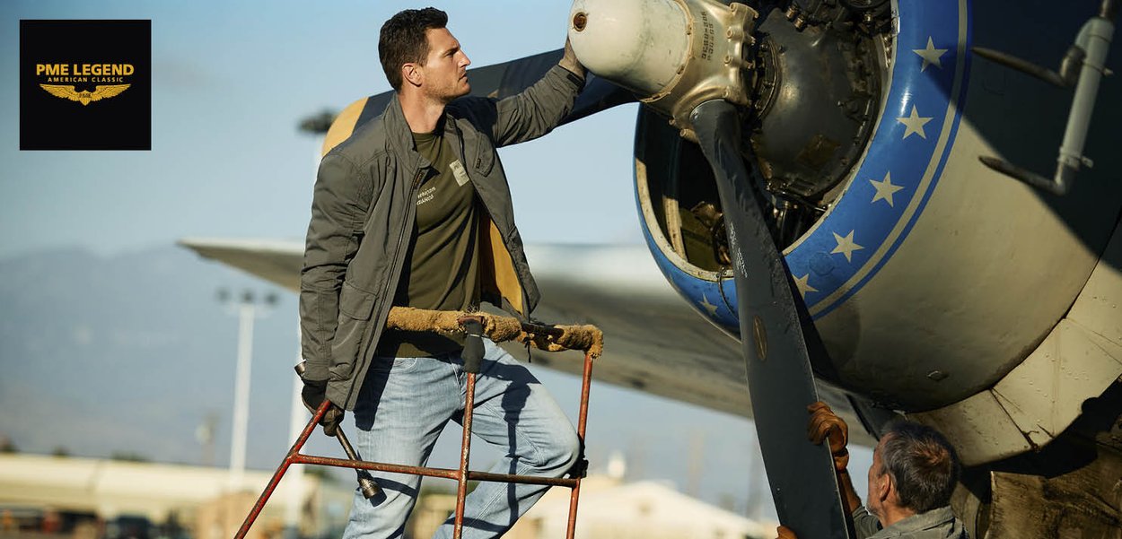Mann in Freizeitjacke inspiziert im Freien den Motor eines historischen Propellerflugzeugs, steht auf einer Metallleiter, während ein anderer Mann das Flugzeug stützt.