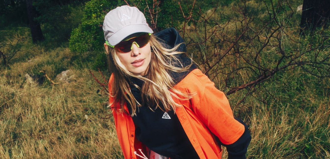 Mujer con gorra blanca, gafas de sol amarillas, sudadera negra y chaqueta naranja, de pie al aire libre en un área boscosa con césped.