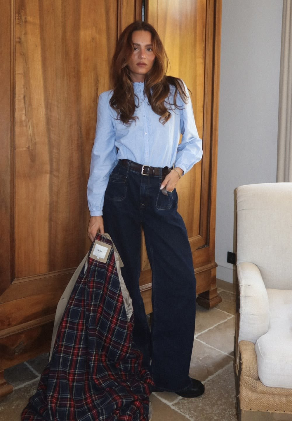 Light blue button-up shirt, dark wide-leg jeans, black belt; holding a plaid patterned coat in navy, red, and white; wood backdrop.