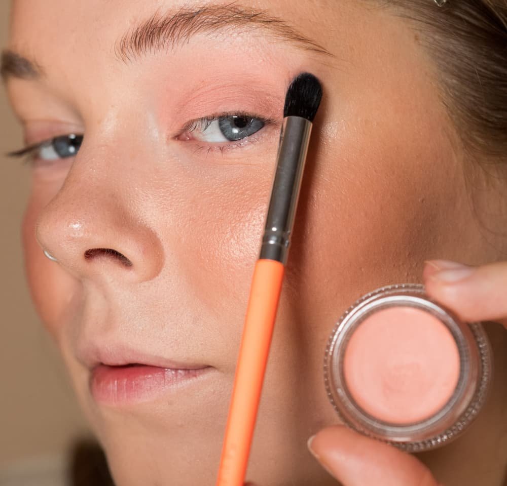 Ferskenfarvet cremeblush i en klar rund beholder, holdt af en hånd. En børste med et orange håndtag påfører blush på kinden af en person.