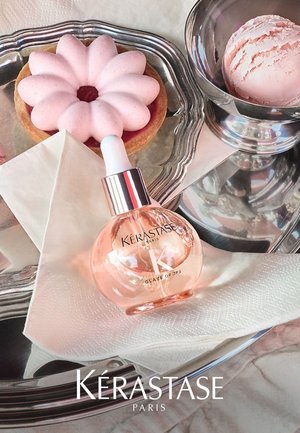 Kérastase Glaze Drops in einer klaren Glasflasche mit Tropfer. Umgeben von einem rosa blütenförmigen Gebäck und einer Tasse pinkem Eis auf einem silbernen Tablett.