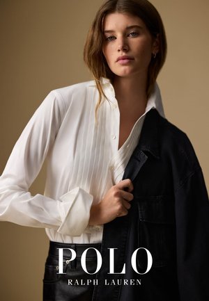 Camicia bianca a maniche lunghe con dettagli plissettati e chiusura a bottoni, sovrapposta a un giubbotto scuro, su uno sfondo neutro.