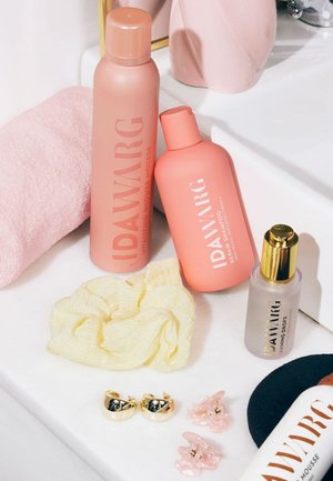 Due bottiglie IDAWARG rosa (shampoo e mousse doccia) accanto a un elastico giallo, orecchini dorati e fermagli per capelli su una superficie bianca.
