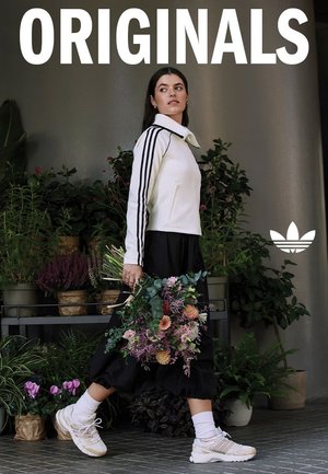 Giacca bianca con zip e strisce nere, gonna nera e sneaker beige. Tiene in mano un bouquet di fiori misti in un ambiente verde e rigoglioso.