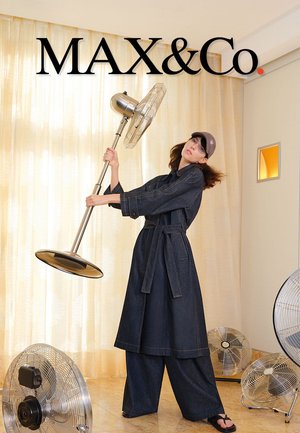 Femme en tenue en denim foncé et casquette tenant un grand ventilateur sur pied, entourée de trois autres ventilateurs dans une pièce lumineuse avec des rideaux transparents.