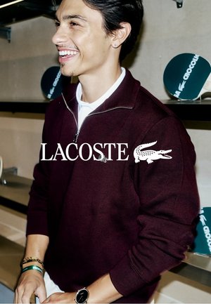 Joven sonriente que lleva un suéter con cremallera Lacoste color granate oscuro con una camisa blanca debajo y un reloj en la muñeca.