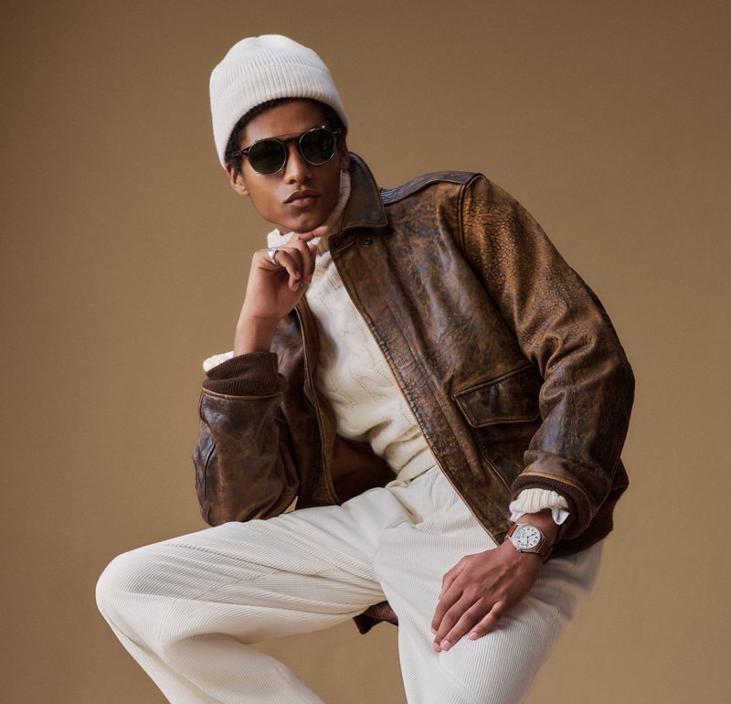 Hombre con gorro blanco, gafas de sol, chaqueta de cuero marrón, suéter blanco, pantalones blancos y reloj de pulsera, posando frente a un fondo beige liso.