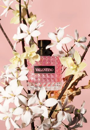 Pinkfarbener Valentino-Parfümflakon, umgeben von weißen und blassgelben Blumen vor einem sanften rosa Hintergrund.