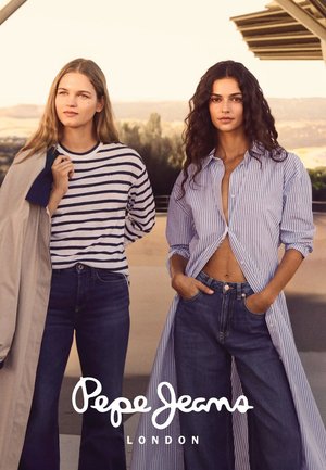 Dos mujeres están al aire libre al atardecer vistiendo ropa vaquera; una lleva una camisa larga de rayas abierta sobre jeans, la otra lleva una camisa de rayas con un abrigo sobre el hombro.