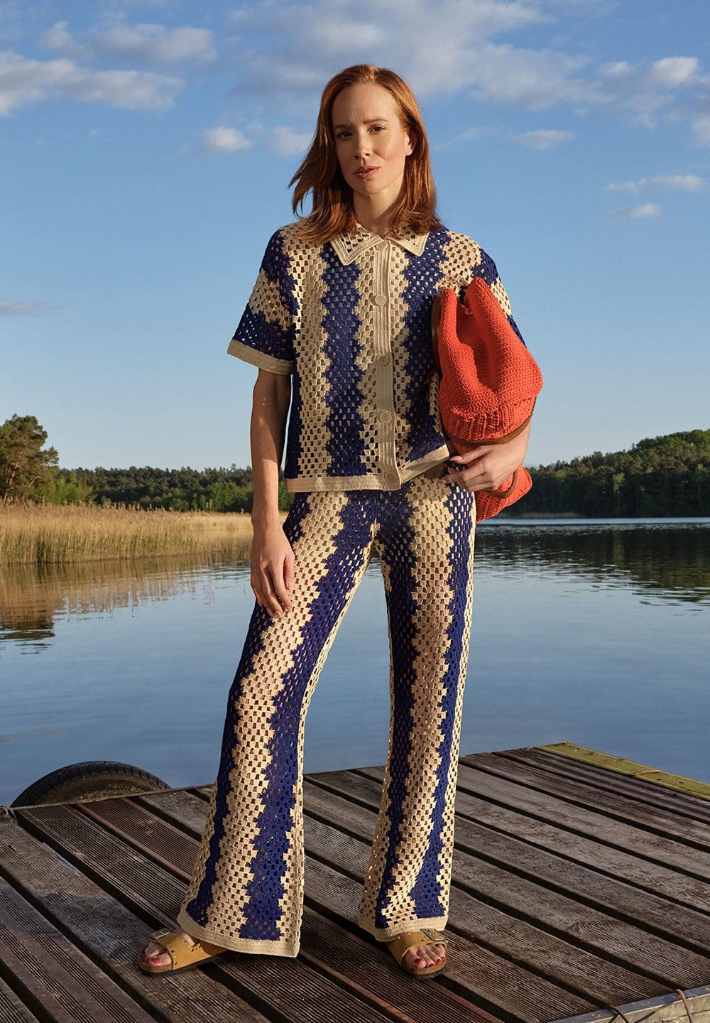 Dva kusový háčkovaný outfit v námořnické modré a béžové barvě se pruhovaným vzorem. Model drží červenou háčkovanou tašku a stojí na dřevěném mole u vody.