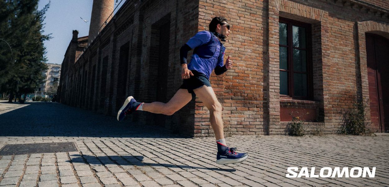 Running shoes with a dark blue upper, white sole, and red accents. The runner wears a short-sleeved shirt and shorts on a cobbled street.