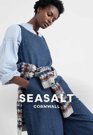 Robe salopette en denim bleu foncé, superposée à une chemise bleu clair avec un col à volants. Pull tricoté multicolore noué autour de la taille.