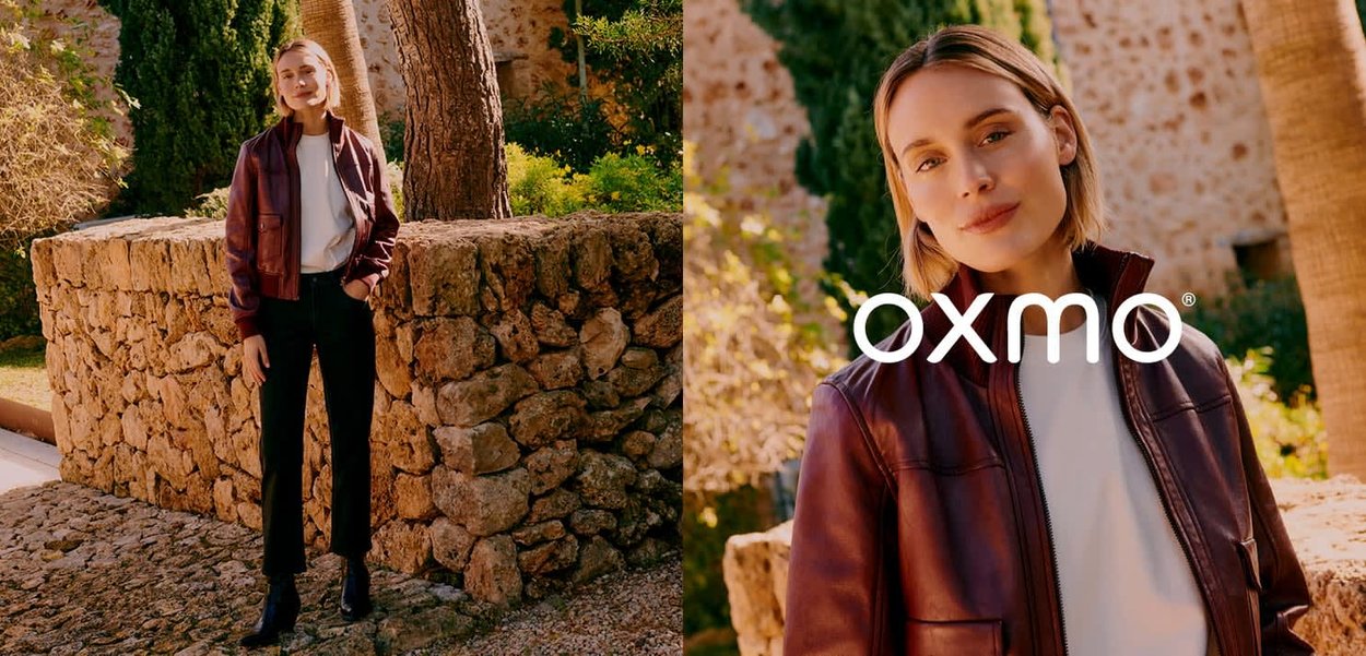 Kvinde iført en bordeaux rød læderjakke, hvid top og sorte bukser, stående på en stensti ved siden af en rustik stenmur og træer.