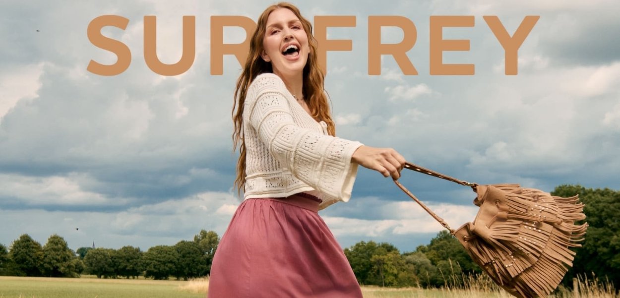 Cotton-blend white cropped sweater with bell sleeves and a pink flowing skirt, holding a fringed tan handbag against a cloudy sky background.
