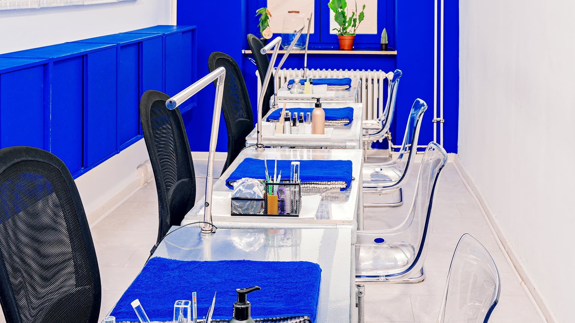 Nagelsalon met doorzichtige acryStoelen en glazen tafels. Blauwe handdoeken en verschillende nagelproducten netjes gerangschikt. Witte muren en blauwe accenten.