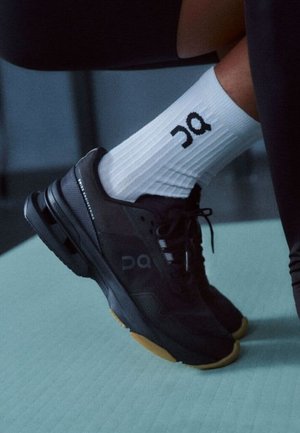 Scarpe da ginnastica nere con suole ammortizzate, indossate con calzini bianchi che presentano un logo "On" nero, mostrate su un pavimento azzurro chiaro.