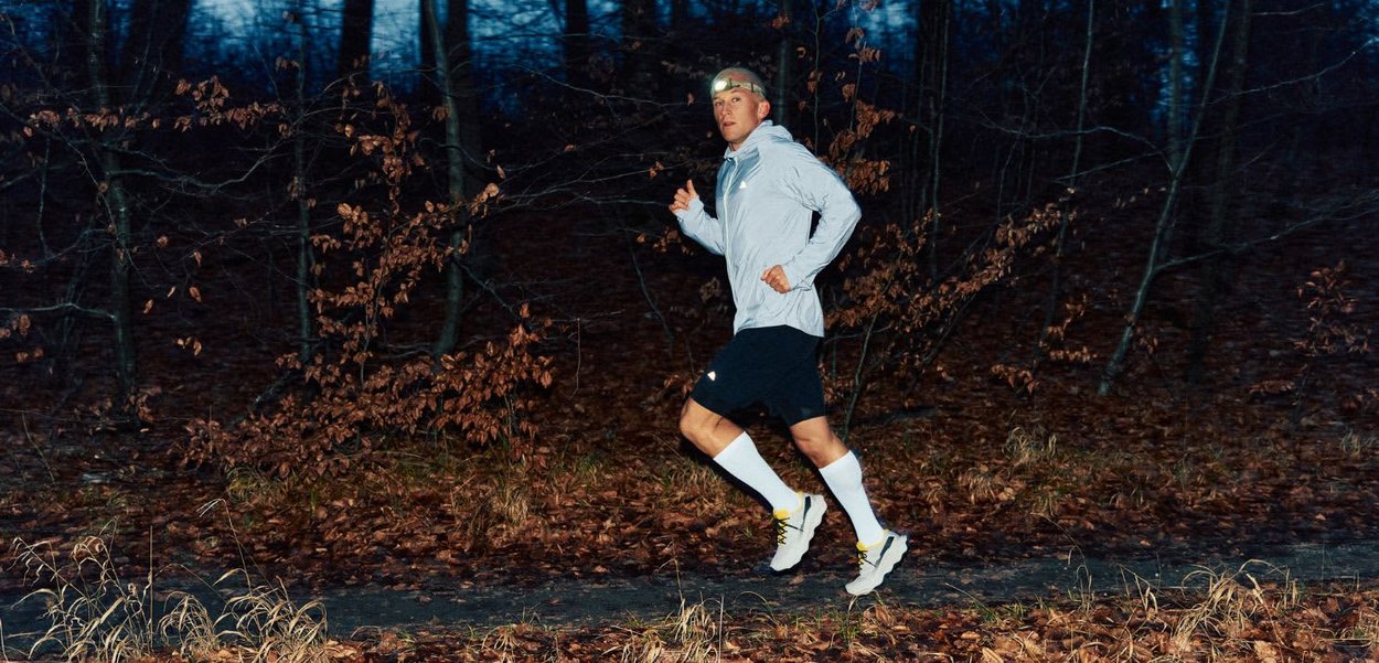 Mężczyzna biegnący leśną ścieżką o zmierzchu, ubrany w jasnoszarą kurtkę, czarne szorty, białe skarpety do kolan oraz czołówkę.