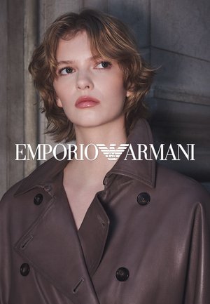 Trench coat de cuero marrón con amplios solapas y cierre de botones dobles. La modelo tiene el cabello corto y ondulado. Marca visible en la parte inferior.