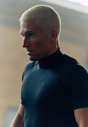 Fit persoon met kort blond haar, gekleed in een zwart sportshirt met hoge hals, gericht en geconcentreerd kijkend naar links in een zacht verlichte binnenruimte.