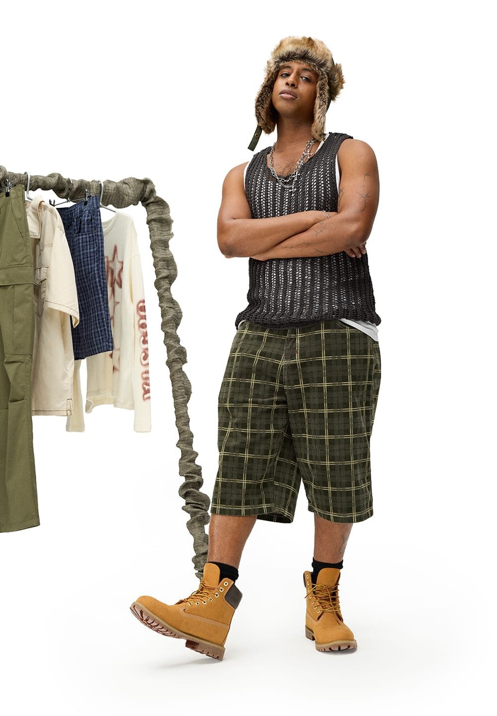 Zwarte gebreide tanktop, groene geruite shorts en tan werkboots met zwarte accenten. Een hangrek toont verschillende kledingstukken op de achtergrond.
