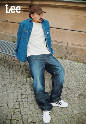 Veste en denim bleue avec poches poitrine sur un t-shirt blanc. Jean bleu à coupe décontractée et baskets blanches avec rayures noires. Casquette marron.
