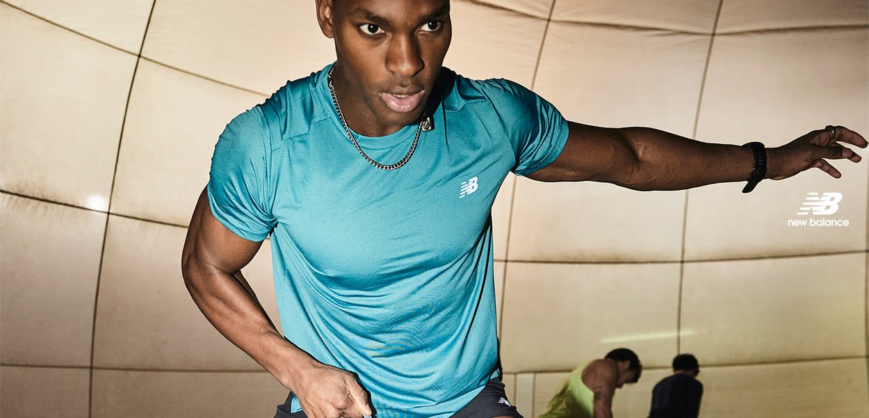 Homme athlétique en maillot bleu New Balance sprintant en intérieur, avec deux autres personnes en arrière-plan près de panneaux muraux courbes.