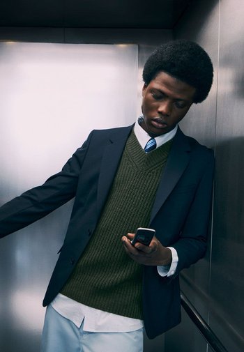 Persona in un completo scuro con un gilet di maglia verde sopra una camicia bianca, che tiene un telefono in mano, appoggiato a un muro dell'ascensore.