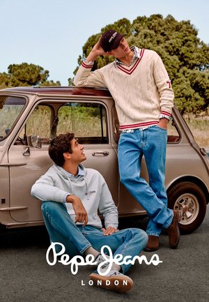 Two young men in casual jeans and sweaters pose near a beige vintage car on a road with trees in the background.