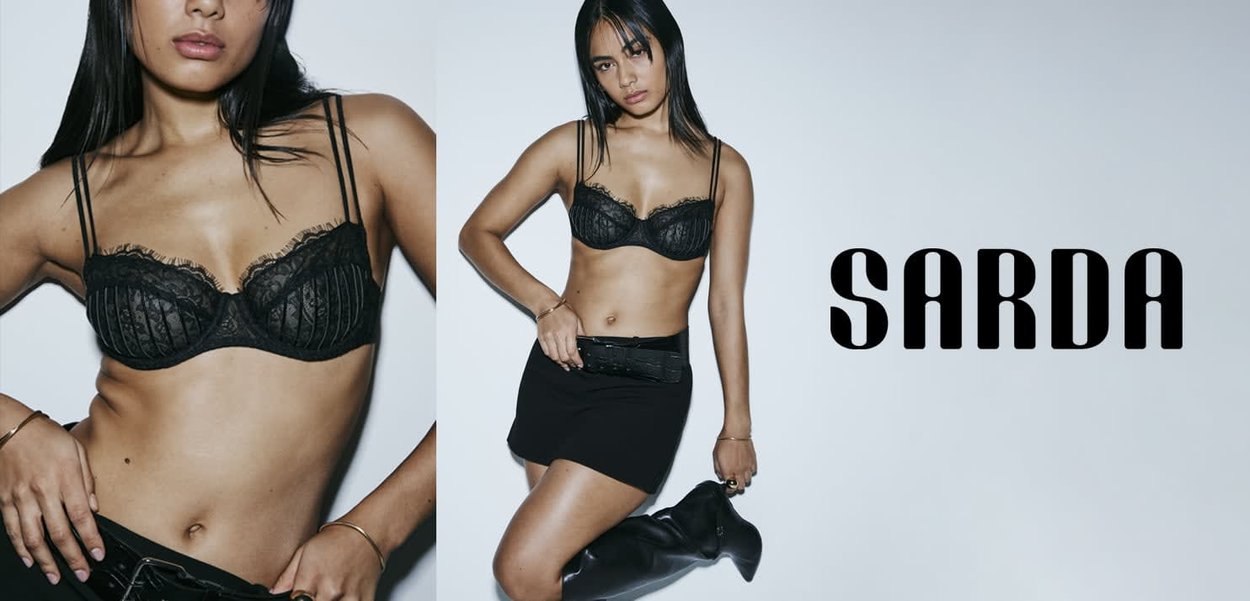 Black lace bra with underwire, featuring vertical pleats and an intricate design. Model wears a black skirt and knee-high boots. Brand logo "SARDA."