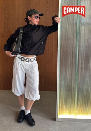 Person in schwarzer, verkürzter Jacke, weißen, weiten Hosen, schwarzen Schuhen, Sonnenbrille und Mütze, die sich an einem strukturierten Glaspaneel mit "CAMPER"-Logo lehnt.