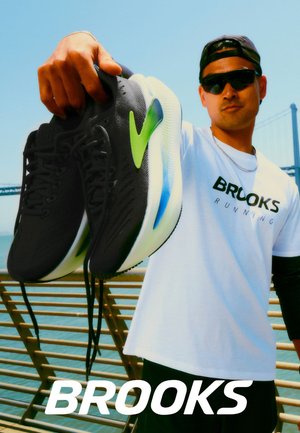 Schwarze Laufschuhe mit grünem Akzent, mit gepolsterter Sohle und strukturierter Oberfläche. Präsentiert von einer Person, die ein weißes Brooks Running Shirt trägt.