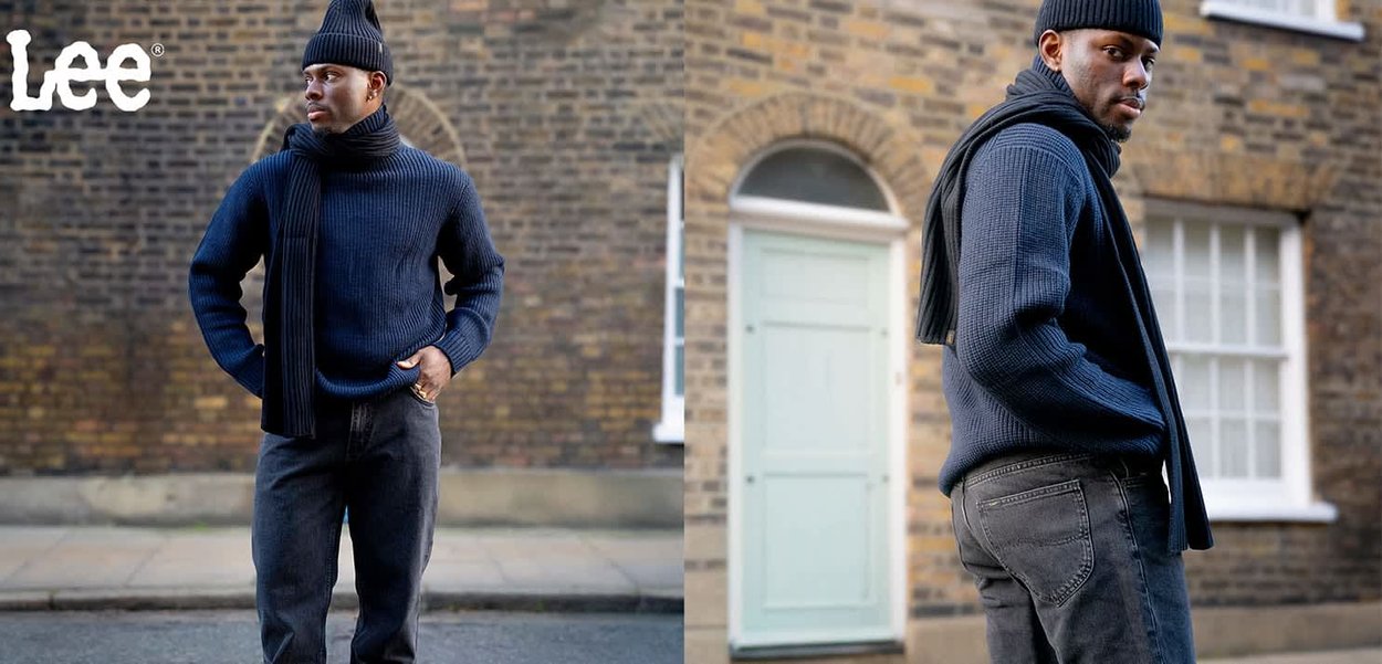 Marinblå stickad tröja med hög krage, kombinerad med en svart halsduk och mörkgrå smala jeans. Bakgrunden visar en strukturerad tegelvägg och en dörr.