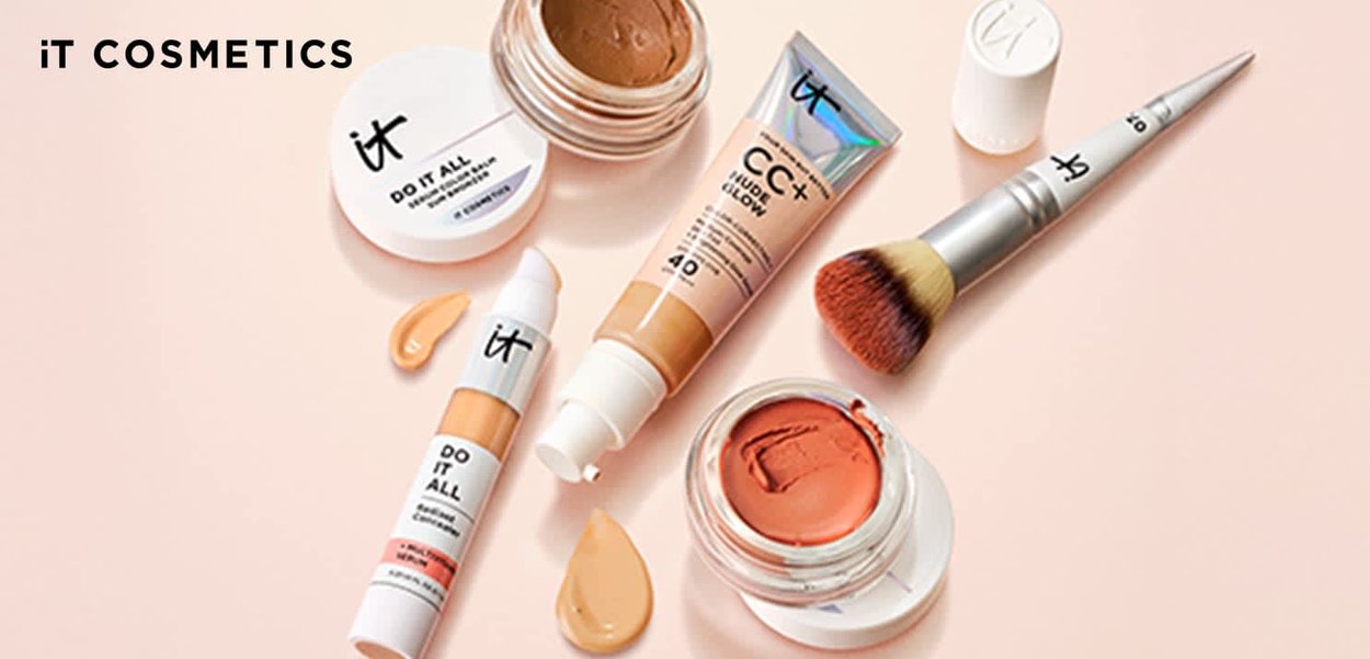 Verschiedene IT Cosmetics Make-up-Produkte, darunter CC-Creme, Concealer, Creme-Rouge, Cremedose und ein Make-up-Pinsel auf hellem Pfirsichhintergrund.