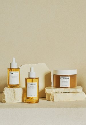 Three skincare products on textured, stacked stone: two amber glass droppers labeled "Centella" and one round brown jar with a white lid.