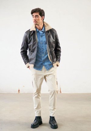 Brown leather jacket with shearling collar, blue denim shirt underneath, light beige cargo pants, and black lace-up boots.