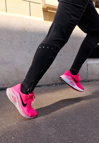 Sneakers Nike rosa con lacci neri, caratterizzati da accenti bianchi e una suola texturizzata. L'atleta indossa leggings neri con dettagli in zip e cucitura.