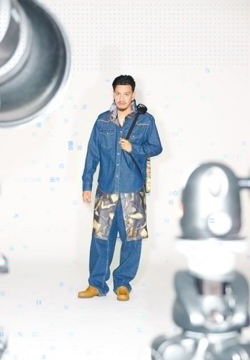 Denimoverhemd met twee borstzakken, camouflage schort en losvallende jeans. Bruine schoenen met gestructureerde zolen. Man staat in een studio-omgeving.