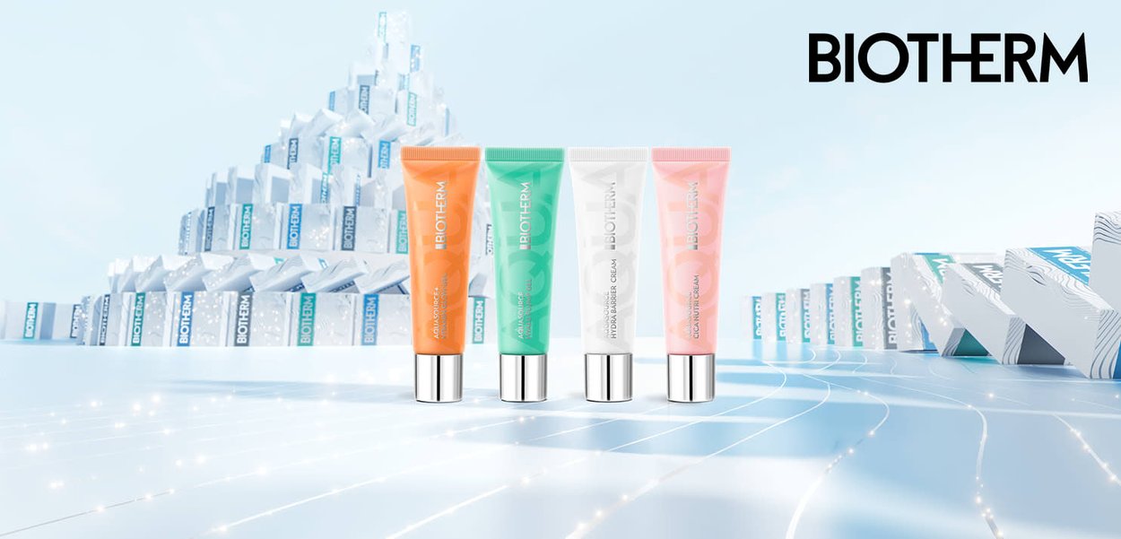 Quattro tubi Biotherm: arancione, verde, bianco e rosa, con tappi argentati. Sullo sfondo si vedono scatole impilate con marchio blu e bianco.