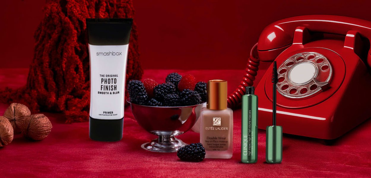 Primers Smashbox dans un tube noir, fond de teint Estee Lauder dans une bouteille en verre, mascara vert Clinique, téléphone vintage rouge, bol en argent de mûres.