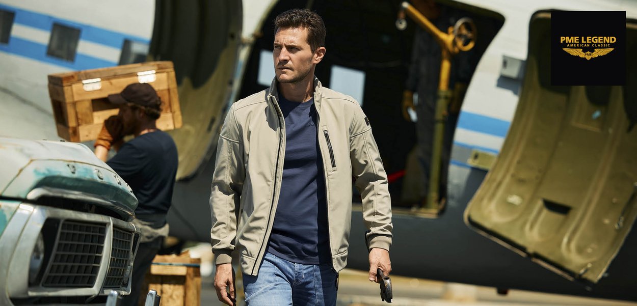 Man in beige jas en blauwe spijkerbroek loopt bij een vliegtuig dat houten kisten lost, houdt een telefoon vast, met het PME Legend-logo op de achtergrond.
