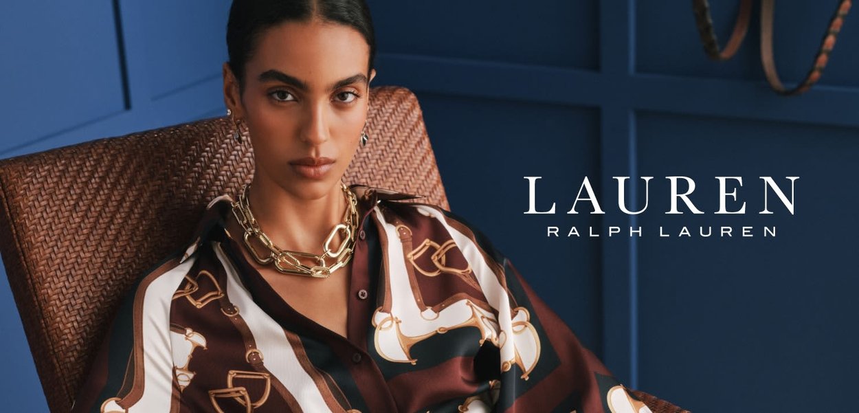 Mujer de cabello oscuro que lleva una blusa de seda con estampado y un collar de cadena de oro, sentada en una silla de mimbre frente a un fondo azul.