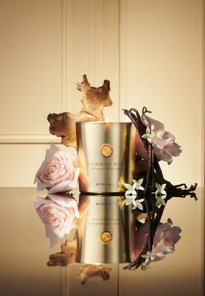 Bocal de bougie en métal doré étiqueté "Cashmere Rose", entouré de roses roses, d'accents en bois et de délicates fleurs blanches sur une surface réfléchissante.