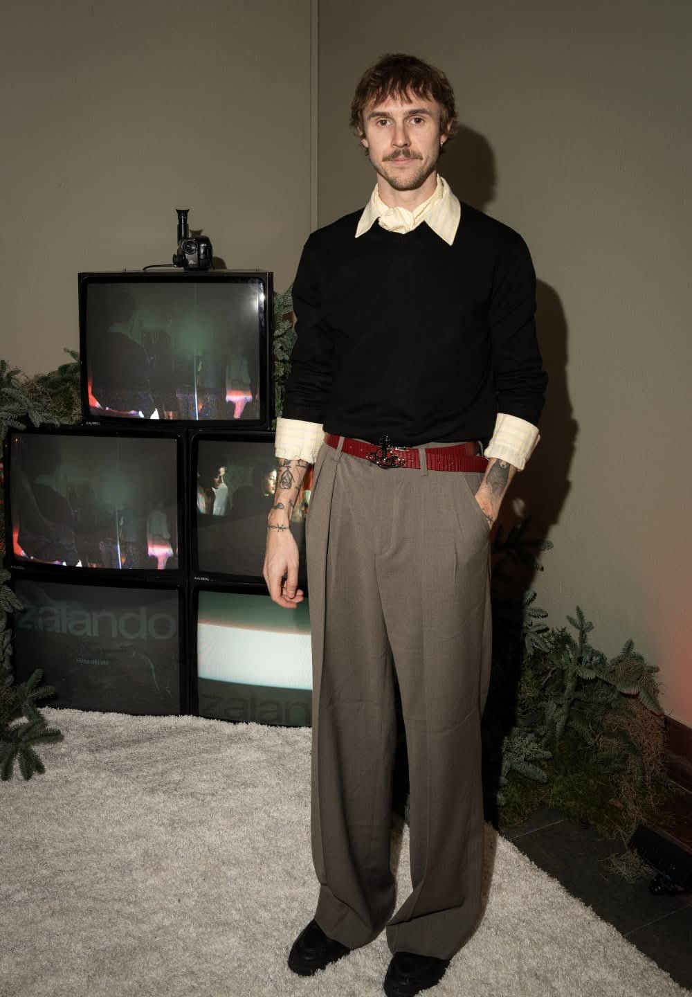 Person iförd en svart tröja, vit skjorta med krage och gråvida byxor med vida ben. Märkbart rött skärp och synliga tatueringar. TV-apparater i bakgrunden.