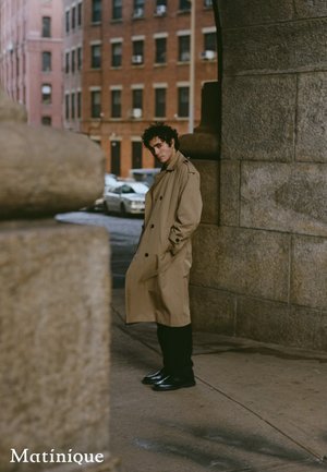 Jeune homme en gabardine beige debout sous une arche en pierre dans une rue de la ville, avec des bâtiments en briques et des voitures garées en arrière-plan.