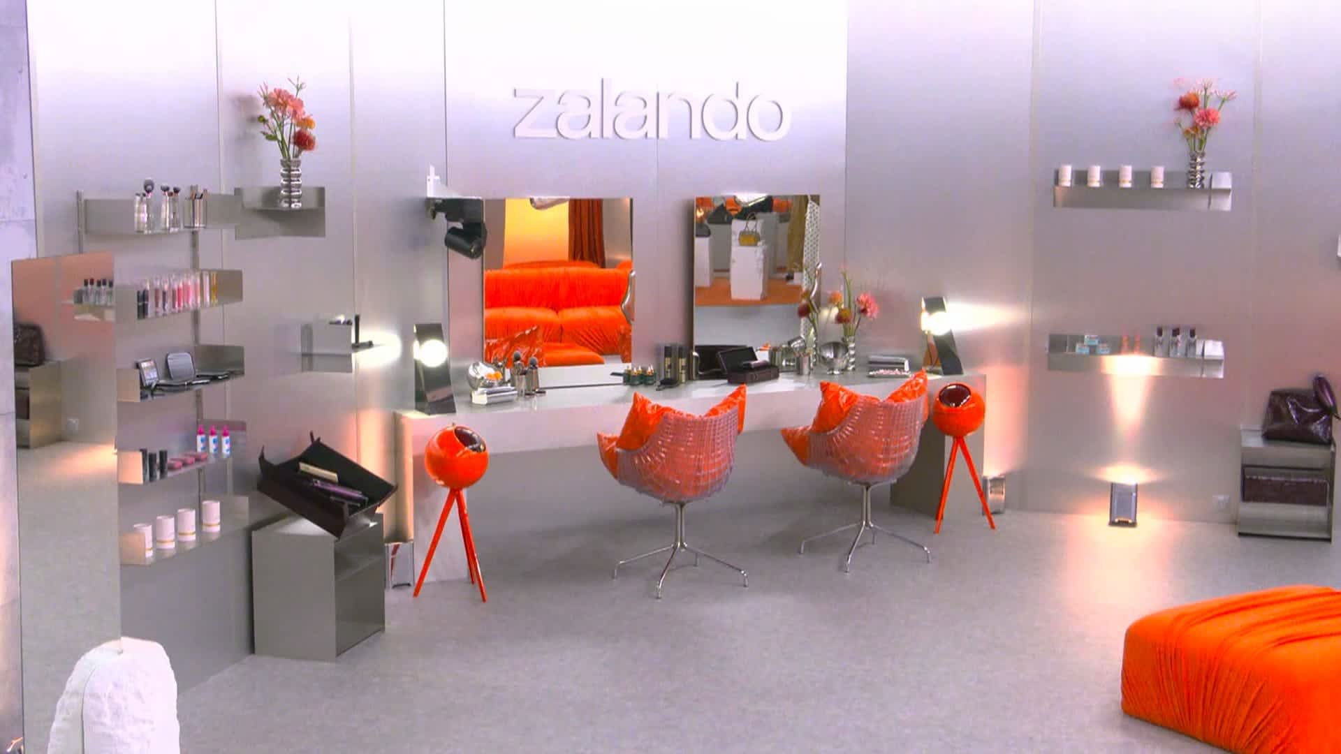 Station de maquillage moderne avec des éléments argentés et orange. Comprend deux chaises oranges, un grand miroir, des étagères et divers articles cosmétiques.