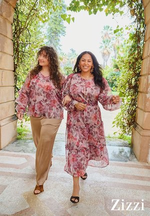 Zwei lächelnde Frauen gehen Arm in Arm unter einem blätterüberdachten Bogen, tragen rosa Blumenmuster-Tops und ein Kleid, im Hintergrund sind Palmen sichtbar.